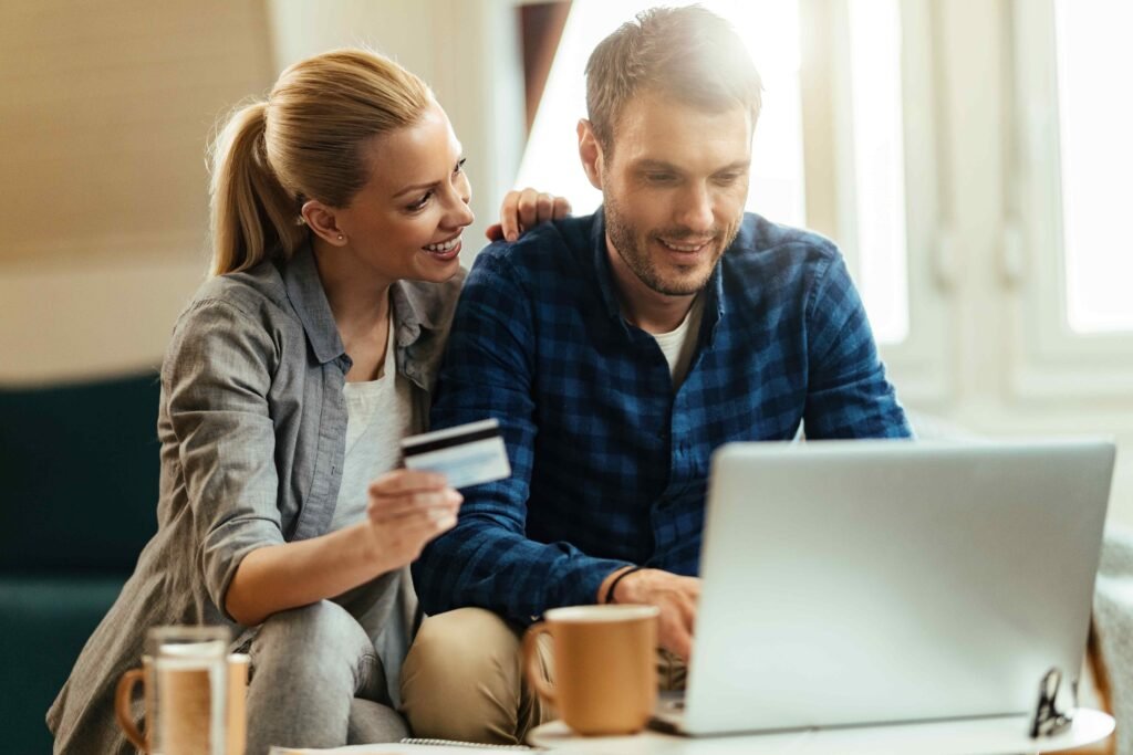 A couple smiles while online shopping at home. The woman holds a credit card, and the man types on a laptop. Coffee mugs are on the table. Cozy atmosphere.