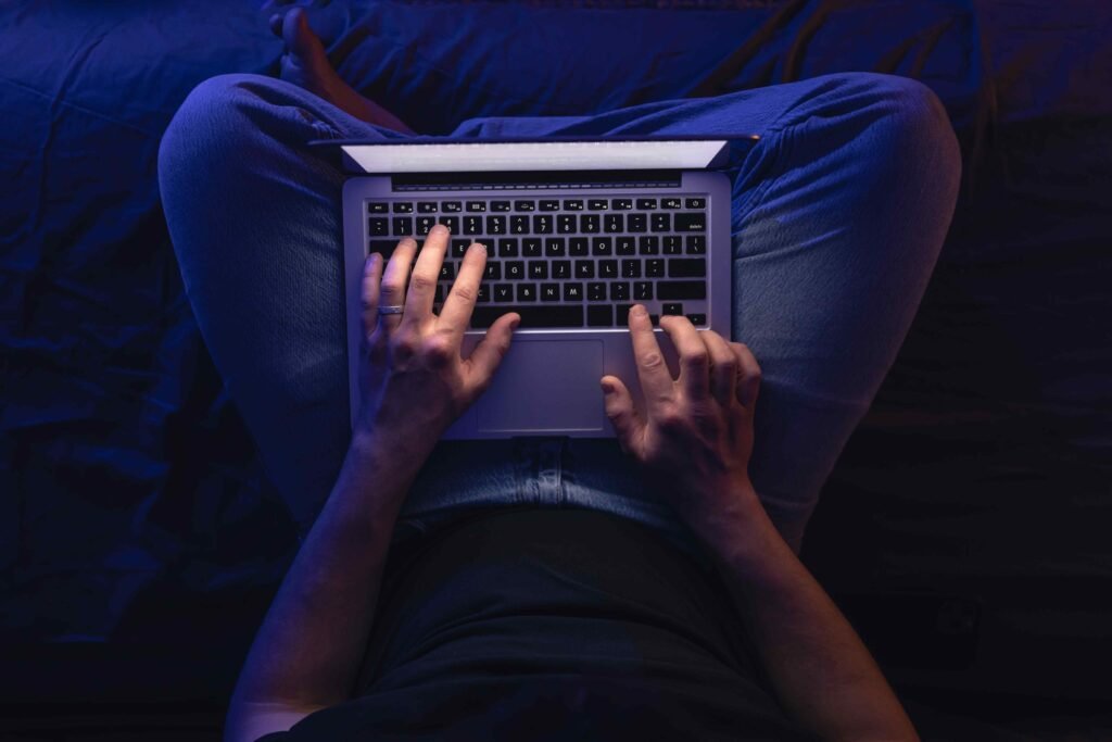 A person types on a laptop while sitting cross-legged on a bed in a dimly lit room, casting a soft glow. The mood is focused and serene.